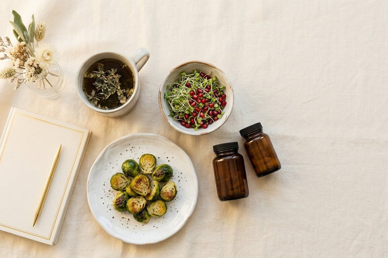 Herbal tea, Brussels sprouts, broccoli sprouts with pomegranate, a journal and amber supplement bottles for natural hormone balance